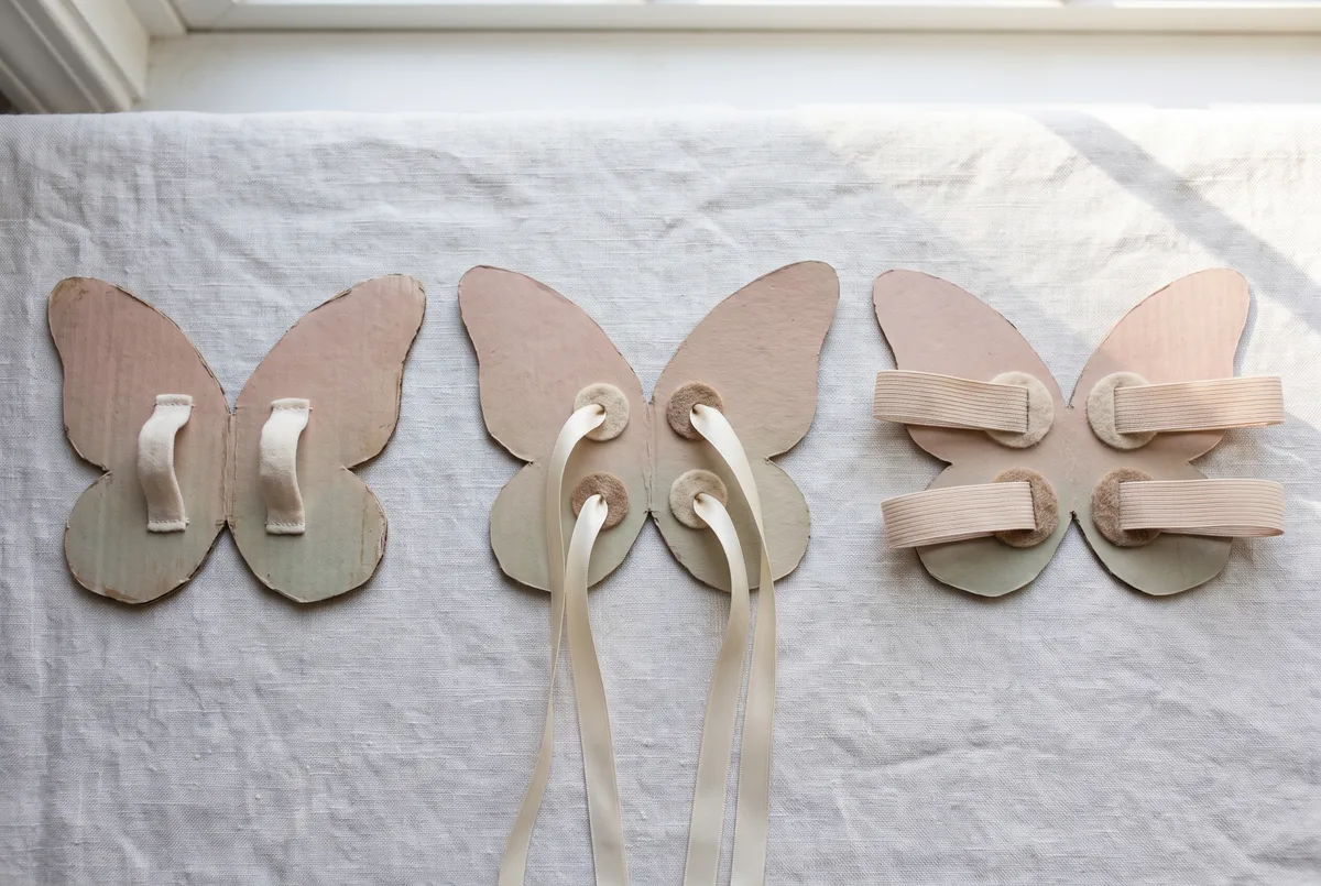 Three fairy wing strap options shown from the back side by side on pale linen: a pair with two soft fabric hand loops, a pair with cream ribbon shoulder loops threaded through felt-reinforced holes, and a pair with wide soft elastic shoulder straps.