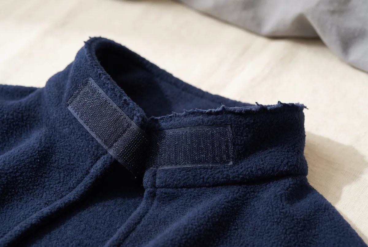 Close detail of a deep navy fleece cloak collar laid flat, showing the folded-over collar band with a wide strip of hook-and-loop tape at each end pulled slightly apart so both the hook and soft loop sides are visible, and the raw unhemmed cut edge of the fleece.