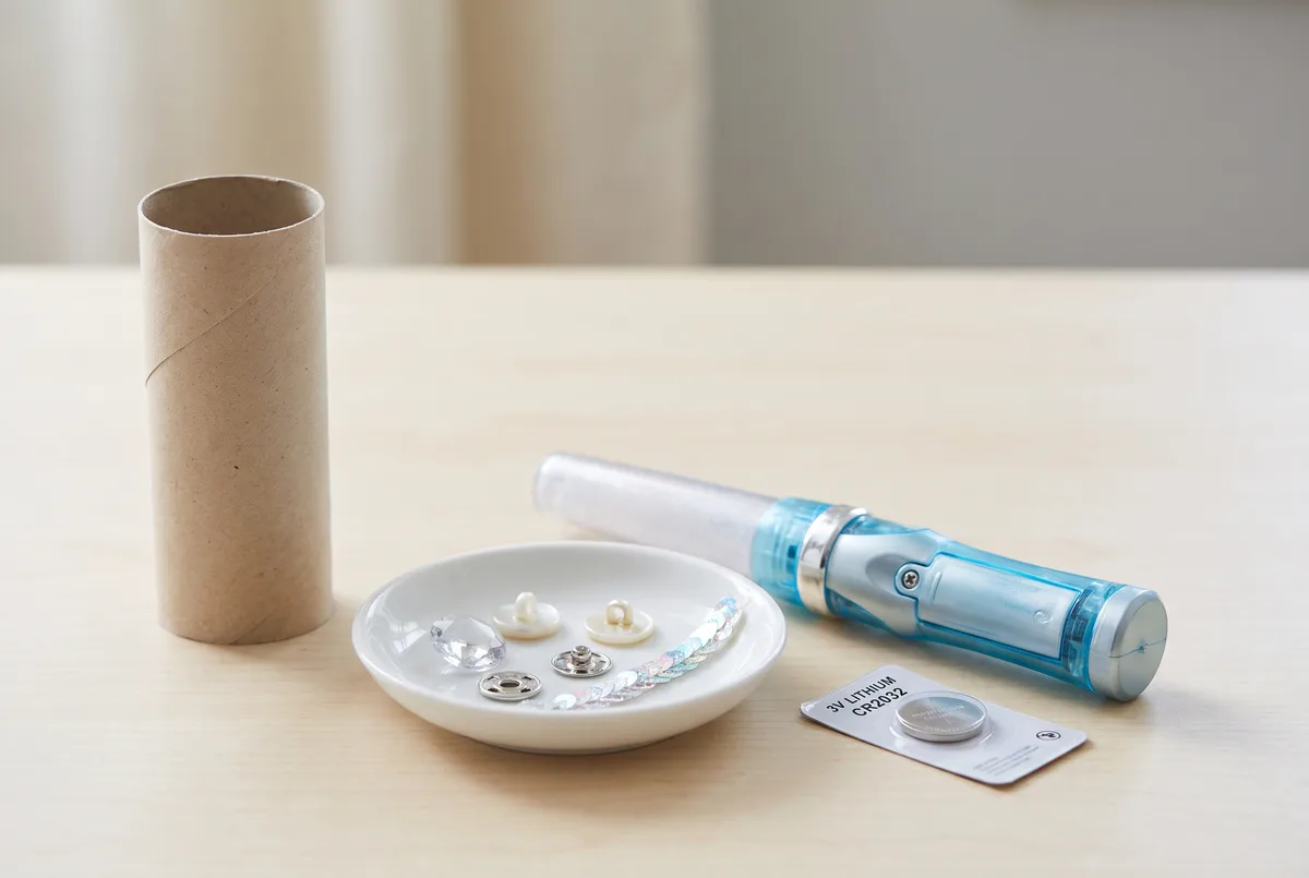 A safety check on a pale wooden table: an empty cardboard tube standing upright, a small dish holding a loose plastic gem, buttons, a snap fastener and sequin trim, a light-up wand with a visible screw-shut battery compartment, and a CR2032 coin battery sealed in its blister card.