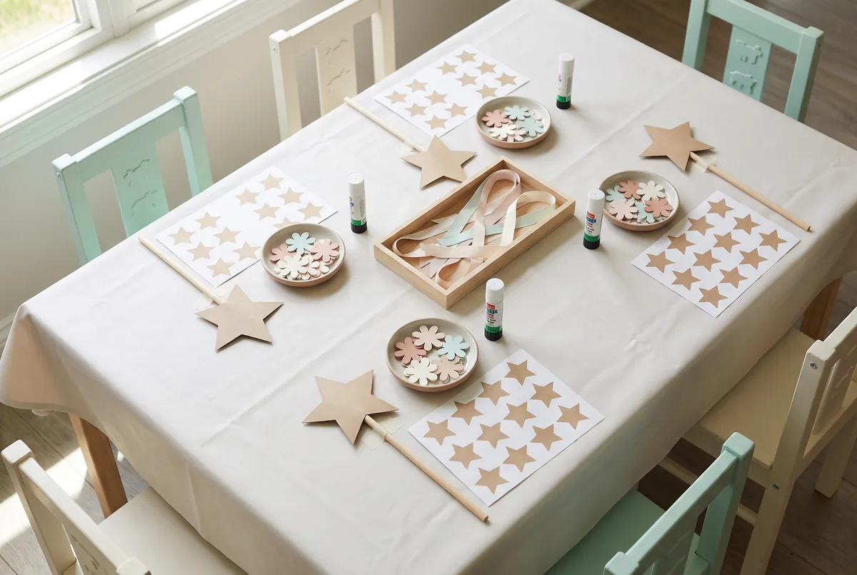 A low children's craft table set for four, each place with a pre-cut card star wand on a wooden dowel, a dish of paper flower shapes, a sheet of star stickers and a glue stick, with a shared tray of ribbon offcuts in the middle.