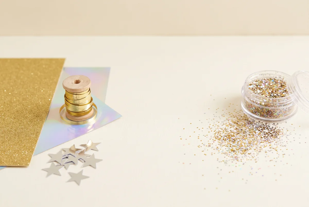 A comparison of party sparkle materials: on the left, neatly arranged gold glitter card, a spool of metallic ribbon, holographic paper and cut foil stars; on the right, set apart, an open pot of loose craft glitter with a scattered spill across the surface.