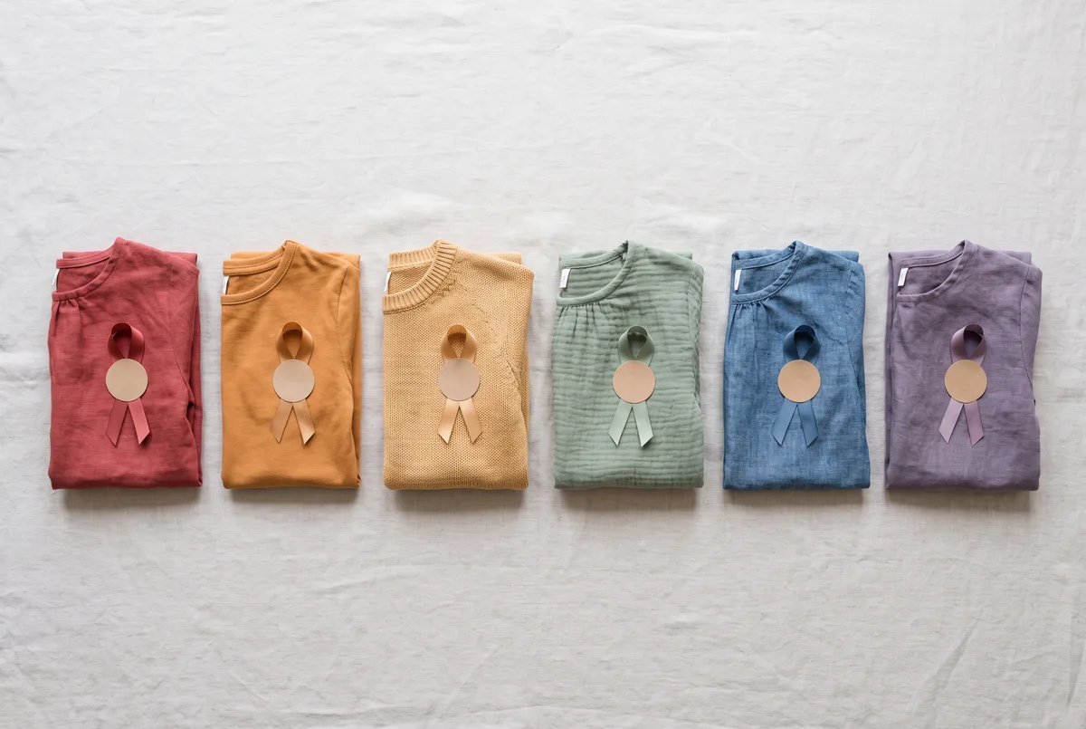 Six children's party outfit sets laid out in a row on pale linen, one per muted rainbow colour — dusty red, warm amber, soft ochre, sage, denim blue and muted violet — each a plain folded top with one matching ribbon and one matching paper badge.