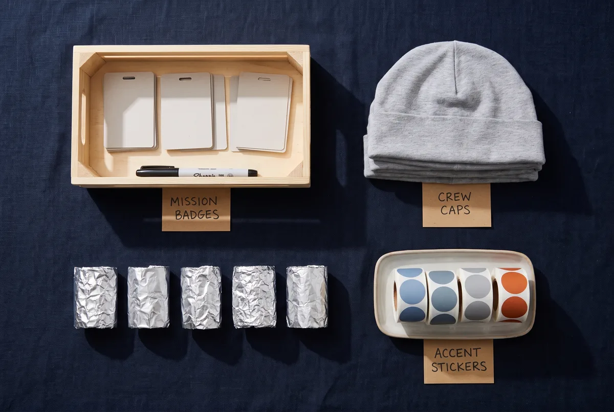 A children's party crew station shot from above on a navy cloth: a crate of blank card mission badges with a marker pen, a row of six silver foil wrist cuffs, a stack of plain grey caps, and a tray of coloured circle stickers, each zone labelled with a small kraft card.