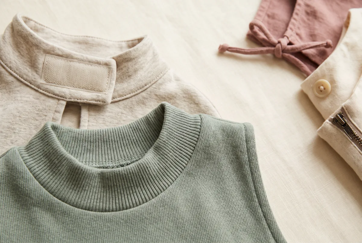 A close comparison of costume closures: on the left in sharp focus a wide soft hook-and-loop tab on a jersey cape collar and a generous stretchy pull-on neck opening on a knit tabard; on the right in softer focus a narrow cord tie, a small hard button, and a back zip pull.