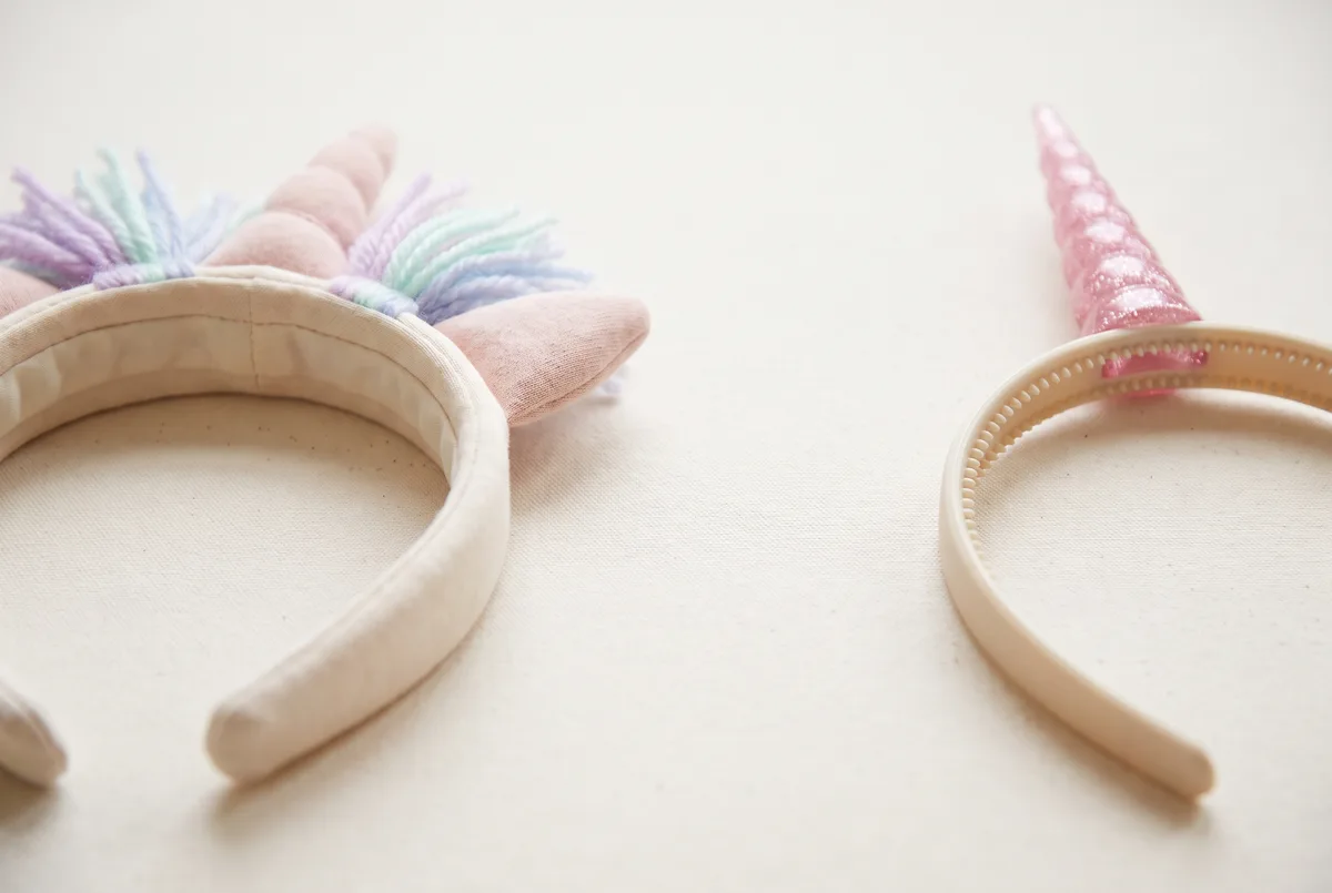 Two unicorn horn headbands side by side for comparison: on the left a wide fabric-wrapped padded band with a short soft stuffed horn and a pastel yarn mane; on the right a narrow bare plastic band with visible gripping teeth along the underside and a tall rigid glittery horn.